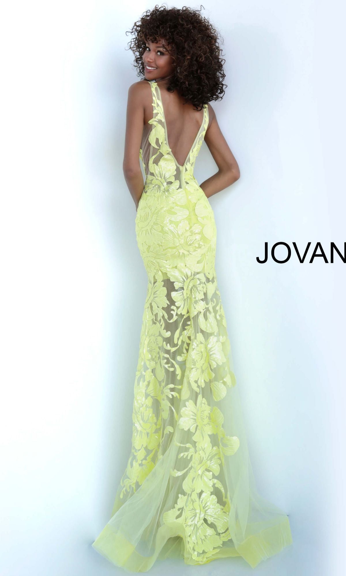 A woman with curly hair models the Jovani Embroidered Long Prom Dress with Sequins, a sheer yellow gown featuring floral embroidery and a deep V-back, posing against a plain background with partial